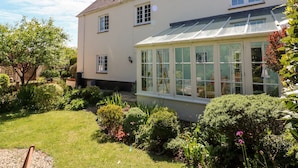 Une maison à deux étages dotée d’une véranda, entourée d’un jardin bien entretenu où poussent différentes espèces de plantes et où s’étend une pelouse.