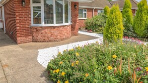A red brick house with a white window frame, a curved driveway, and a garden with various plants and flowers. - Broadway Cottage (Lincoln)