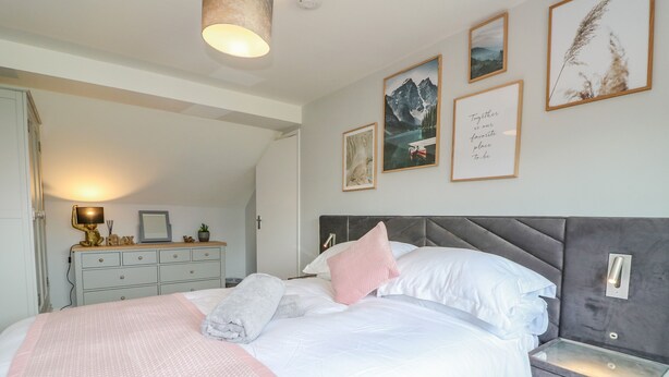 A neatly made bed with pillows and a blanket, a bedside table with a lamp, and framed pictures on the wall. - Broadway Cottage (Lincoln)