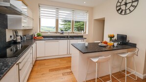 A modern kitchen with white cabinets, a black countertop, and a large window with blinds. - Aislaby Hall (Whitby)