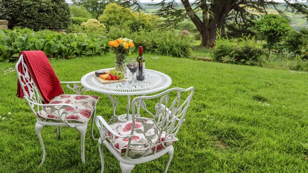 A round table with a white metal frame, two chairs with floral upholstery, and a red cloth draped over one chair. A bottle of wine, a glass, and a fruit platter are on the table. - Aislaby Hall (Whitby)