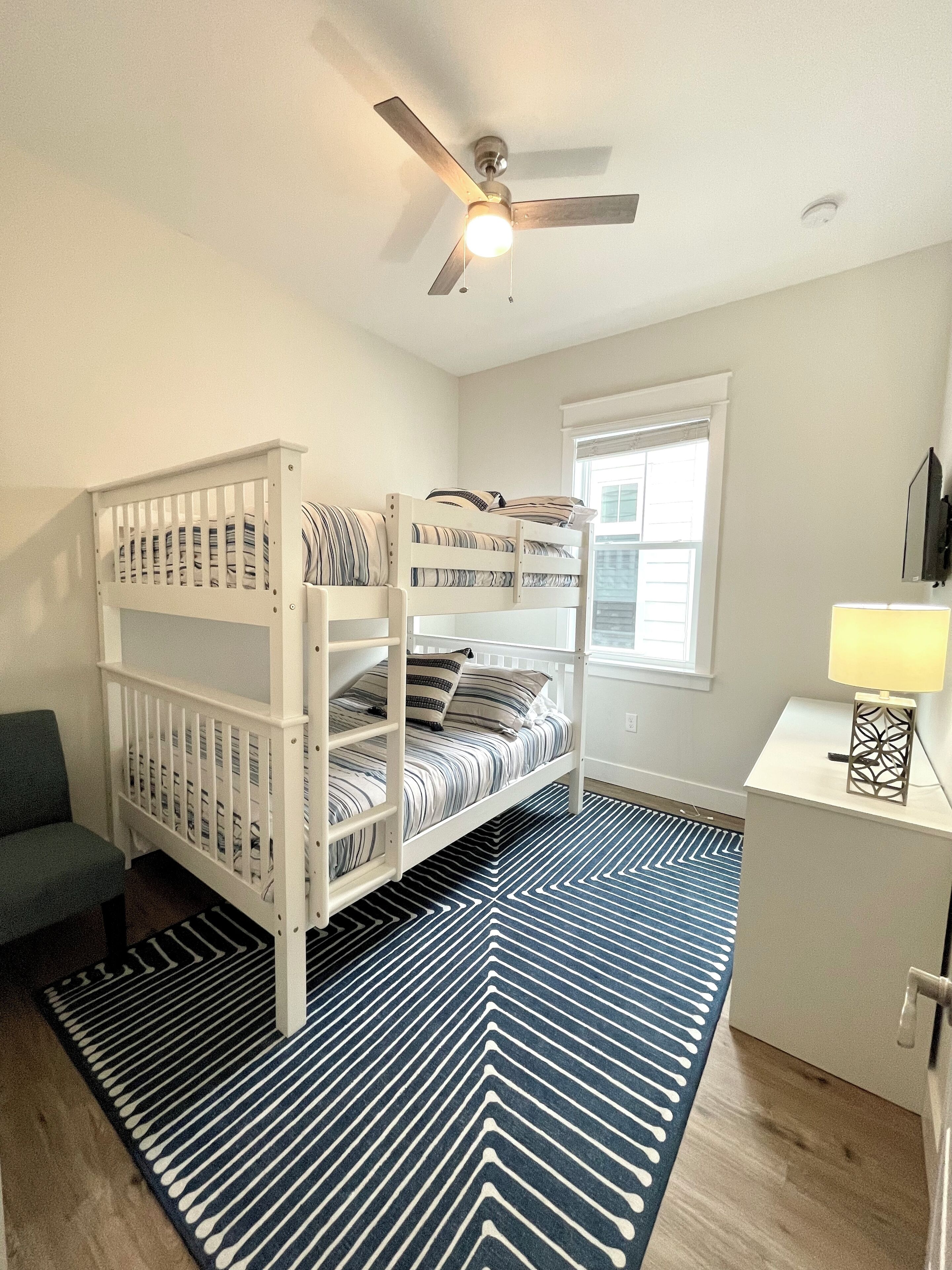 Une chambre avec un lit superposé, un ventilateur de plafond, un bureau avec une lampe et une fenêtre.