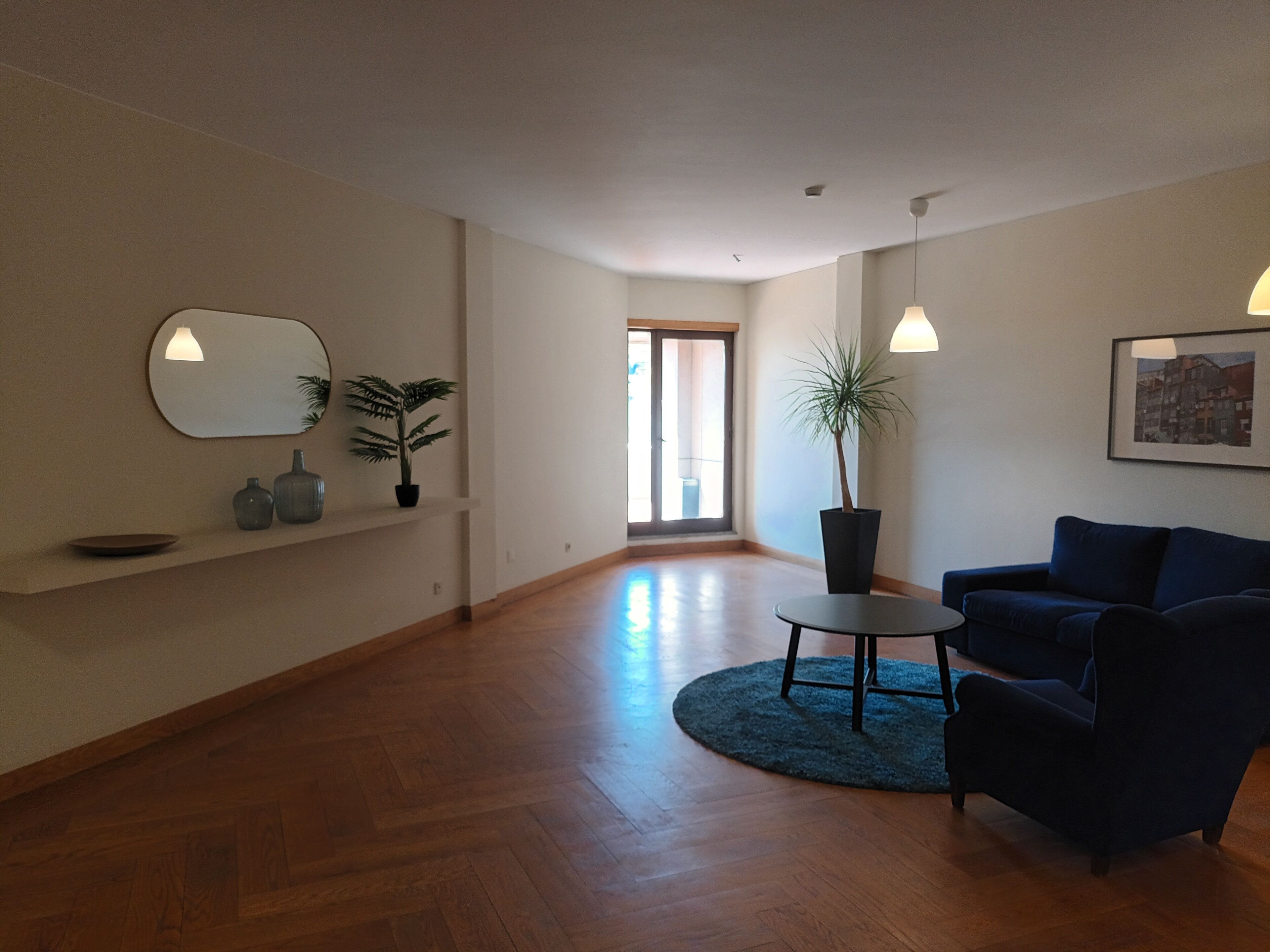 A spacious living room with a wooden floor, a blue sofa, a round coffee table, and a potted plant.