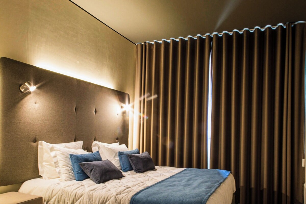 A hotel room with a bed, bedside tables, a headboard, and a window with curtains.