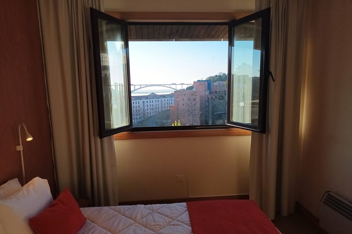 A hotel room with a bed, a window with curtains, and a view of buildings and a bridge.
