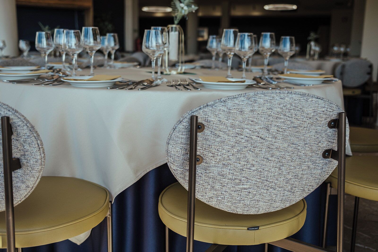 A dining table set with multiple wine glasses, plates, and cutlery. Chairs with round, textured backs are positioned around the table.