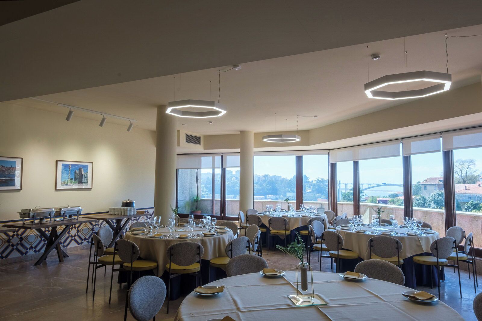 A dining area with round tables set for multiple guests, large windows offering a view of the cityscape, and modern pendant lighting.
