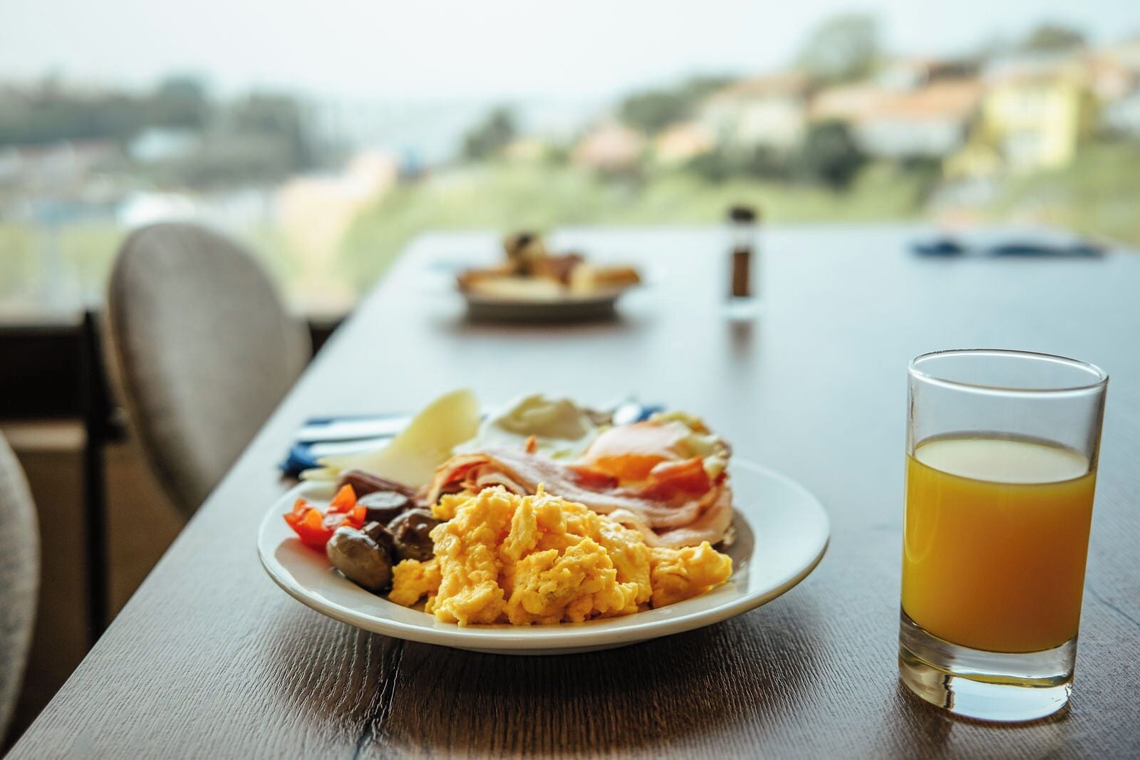 A breakfast plate with scrambled eggs, bacon, and toast, accompanied by a glass of orange juice, on a wooden table with a view of a landscape.