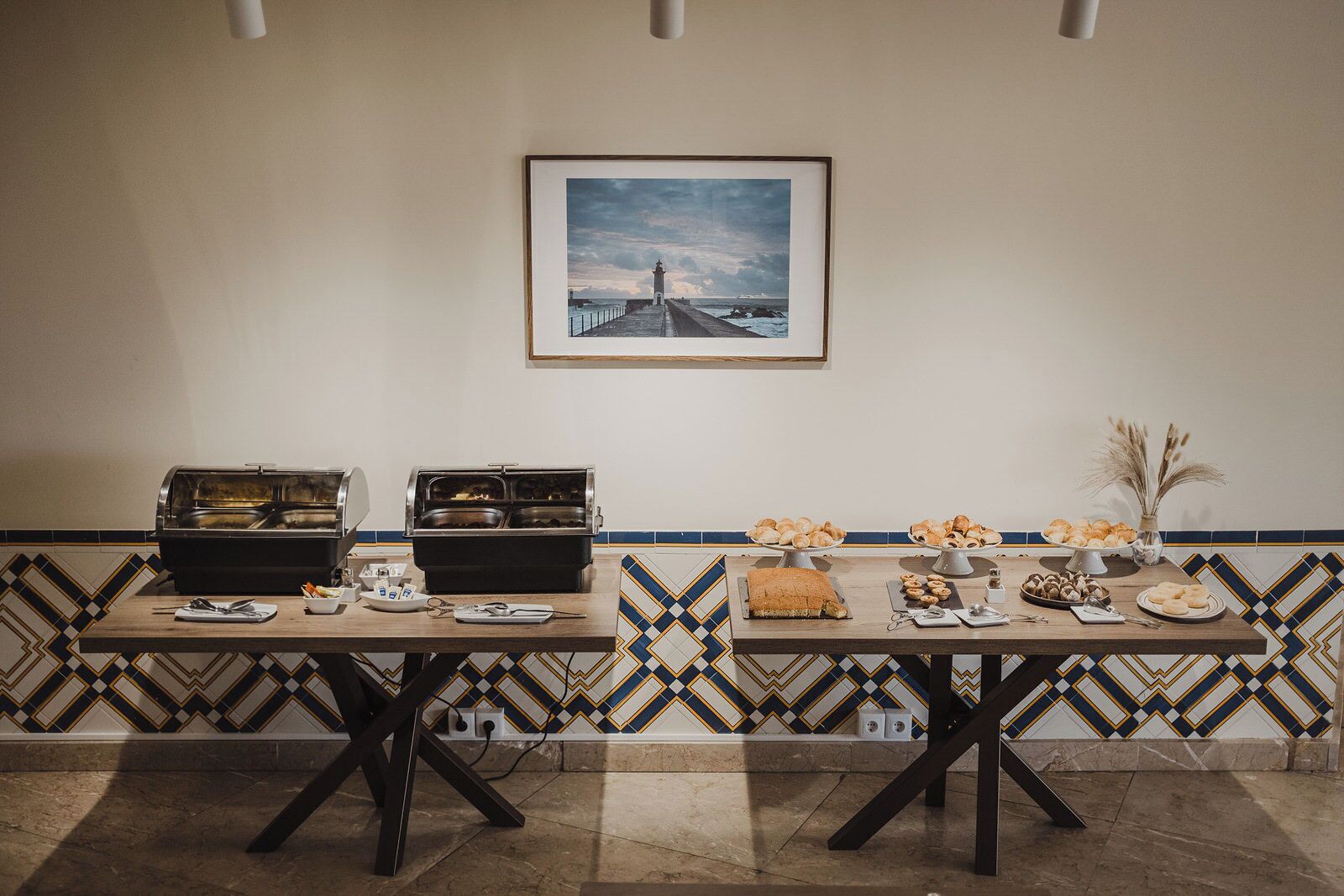 Un'area buffet con due postazioni per cibi caldi, una selezione di dolci e un quadro incorniciato raffigurante un paesaggio costiero.