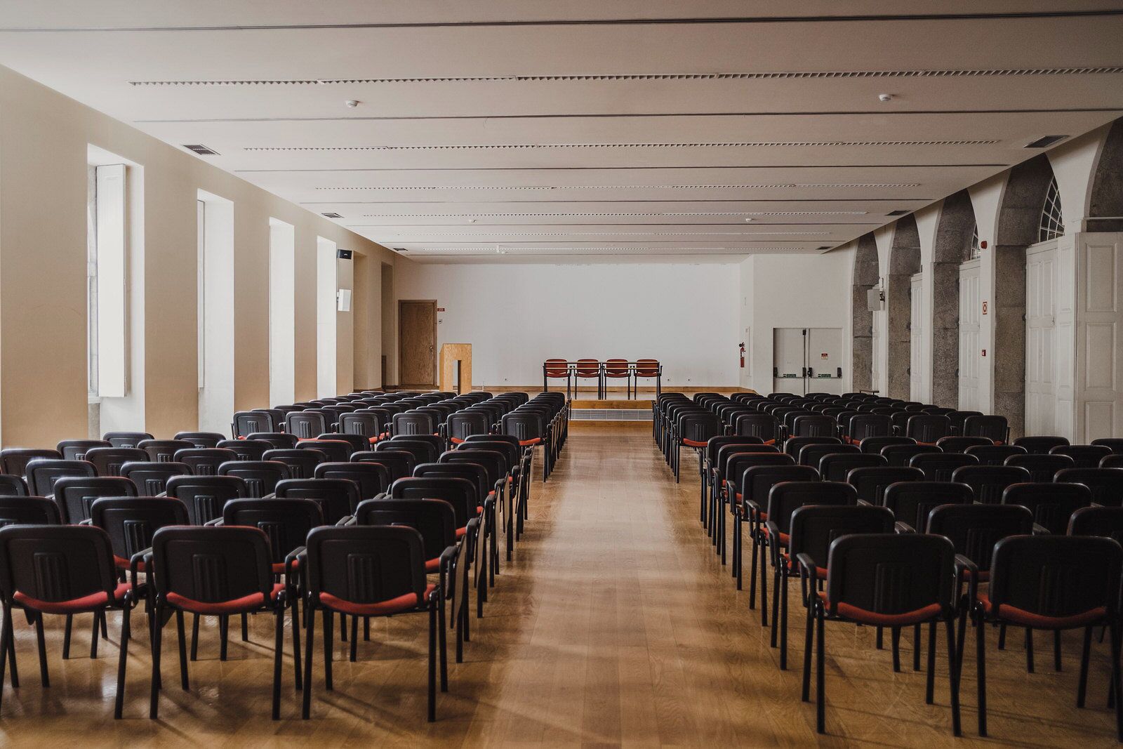Uma sala de conferências com fileiras de cadeiras pretas com almofadas vermelhas, piso de madeira e teto branco com iluminação embutida.