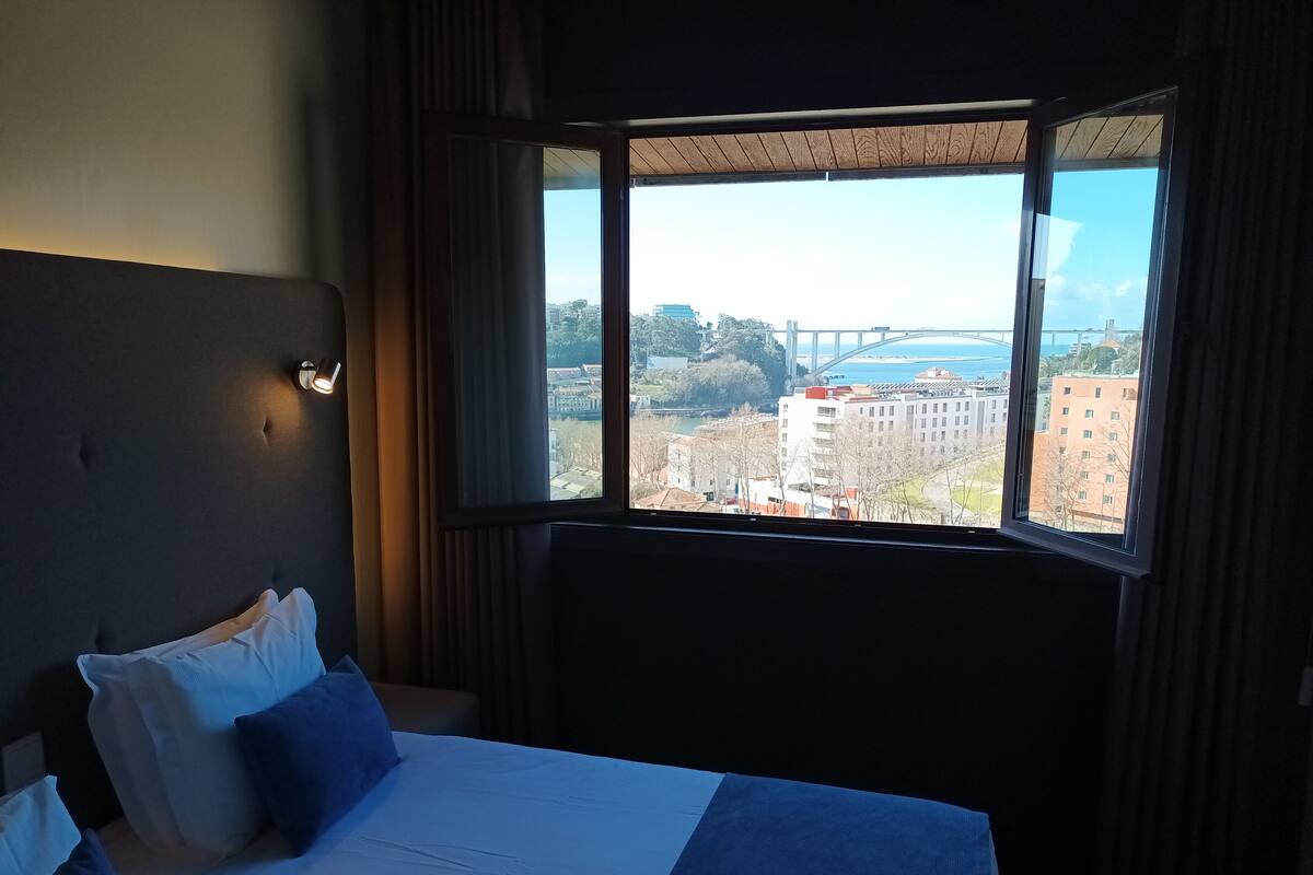 A hotel room with a large bed, a headboard, a nightstand, and a window offering a city view.