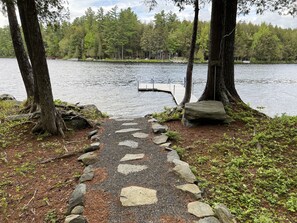 A stone pathway leading to a dock by a lake, surrounded by trees and a small cabin.