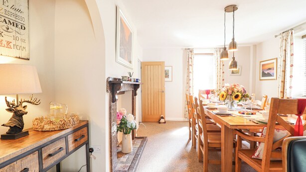 A dining area with a wooden table, chairs, and a chandelier. There is a fireplace with a vase of flowers and a lamp on the left side. - Coed Derw Isaf (Betws-Y-Coed)
