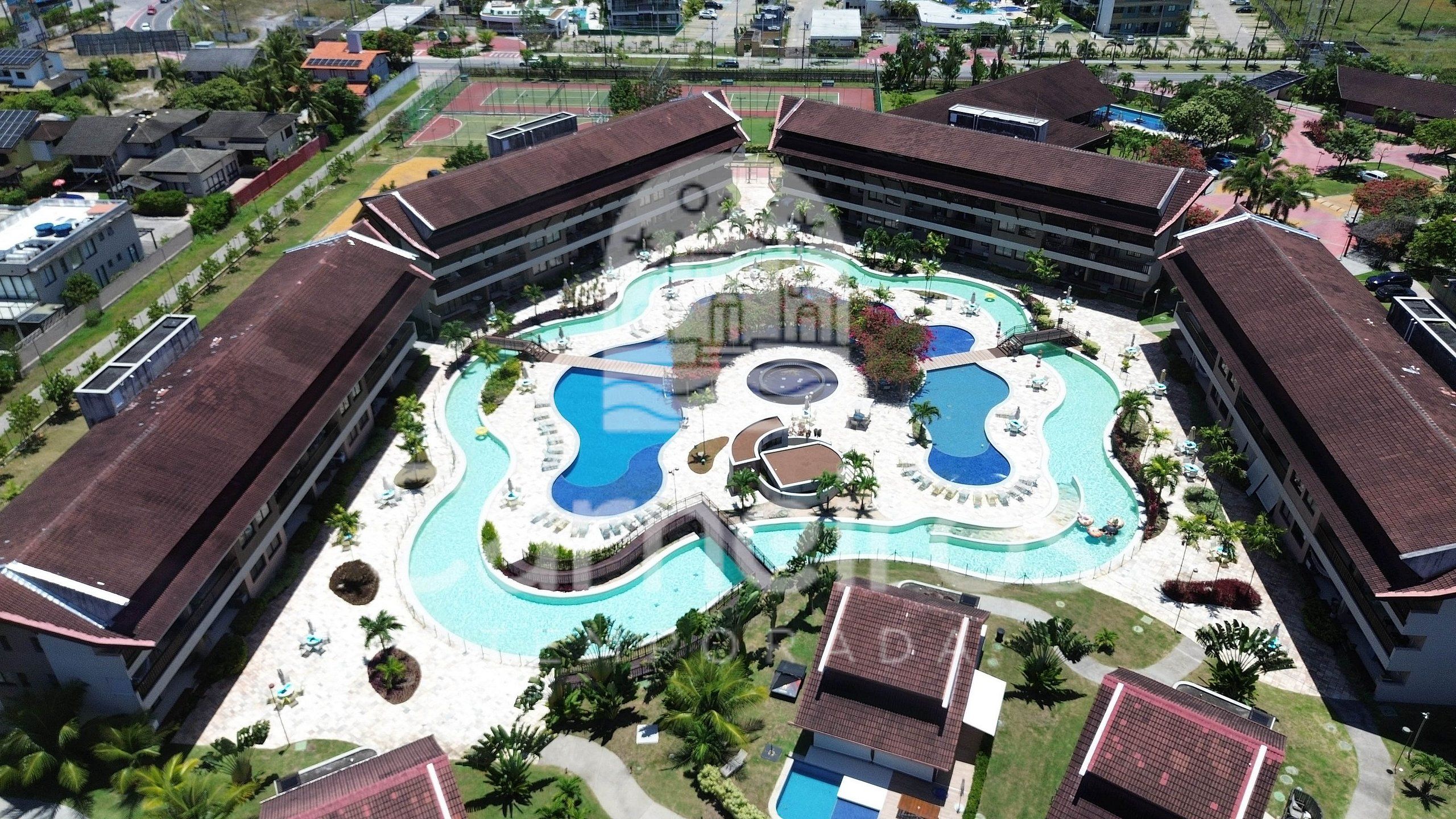 A hotel with a central swimming pool surrounded by multiple smaller pools, a children's play area, and a tennis court.
