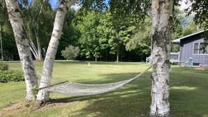 Una hamaca entre dos abedules en un jardín cubierto de hierba, con una casa al fondo.