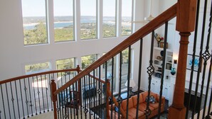 Amplia sala de estar con un gran ventanal que da a un paisaje pintoresco, una escalera de madera y un sofá naranja acogedor.