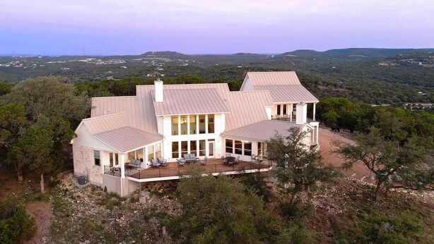 Una casa grande de varios pisos con un amplio balcón y ventanas grandes, rodeada de un bosque denso y colinas.