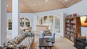 Un salon spacieux avec un plafond en bois, de grandes fenêtres, un canapé, un fauteuil, une table basse et une bibliothèque.