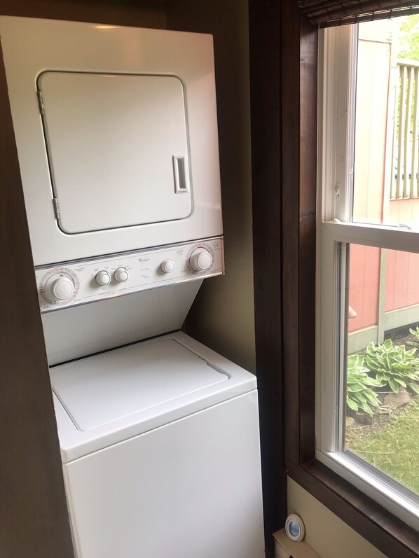 Un lavadero pequeño con lavadora y secadora, una ventana con vista al patio trasero, y una puerta que da a otra habitación.