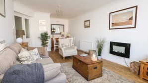 Una sala de estar con sofá, sillón, mesa de centro y chimenea. En el fondo, se ve una zona de comedor con mesa y sillas.