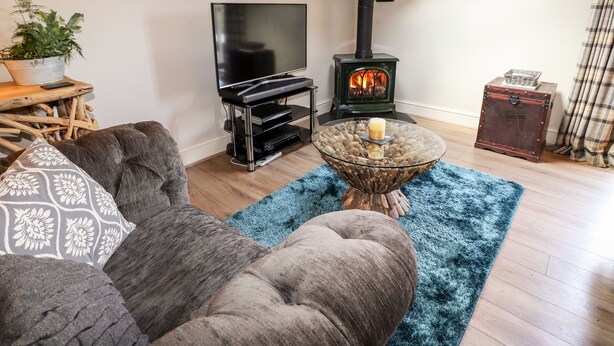 A cozy living room with a flat-screen TV, a black wood-burning stove, a glass coffee table with candles, and a blue rug. - Peony Cottage (Ulverston)