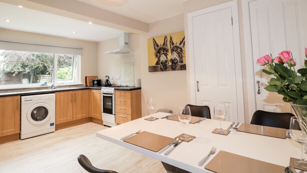 A modern kitchen with wooden cabinets, a built-in oven, and a washing machine. There is a dining area with a table set for two, and a painting of donkeys on the wall. - Alturlie (Inverness)