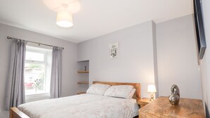 A bedroom with a wooden bed, a bedside table with a lamp, a shelf, and a window with curtains. - Alturlie (Inverness)