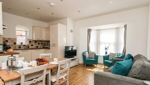 A modern living room with a grey sofa, teal armchairs, and a wooden dining table with chairs. A television is on a stand in the living area, and a kitchen with beige cabinets is visible in the background. - Marina Reach (Weymouth)