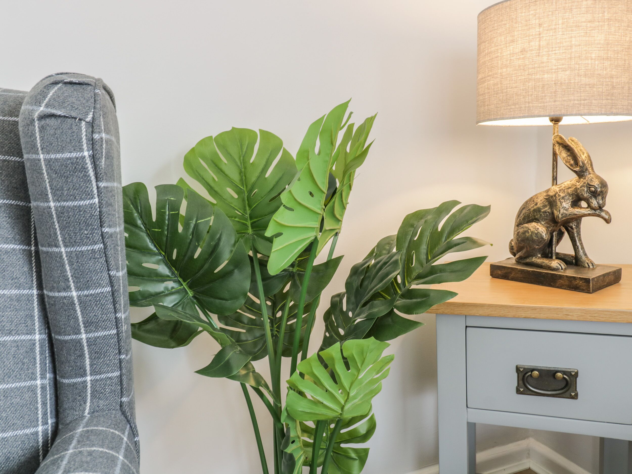 A room with a grey plaid blanket, a green monstera plant, a rabbit statue, and a lamp on a wooden nightstand.