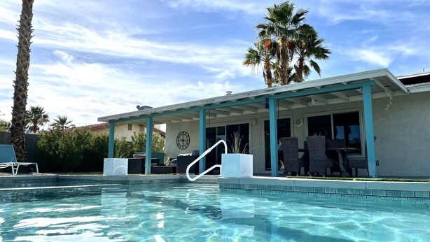 Una piscina con un patio cubierto, palmeras y un reloj en la pared.