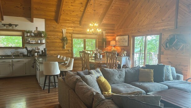A cozy cabin interior with a wooden ceiling, a dining area with chairs and a table, a sofa with cushions, and a kitchen with a sink and cabinets. - The Little Rascals Lake House (Cameron)