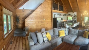 A cozy cabin interior with a large gray sectional sofa, wooden walls, a kitchen area, and a dining table. - The Little Rascals Lake House (Cameron)