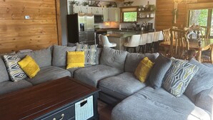 A cozy cabin interior with a large gray sectional sofa, a wooden coffee table, and a kitchen area with a refrigerator, stove, and bar seating. - The Little Rascals Lake House (Cameron)