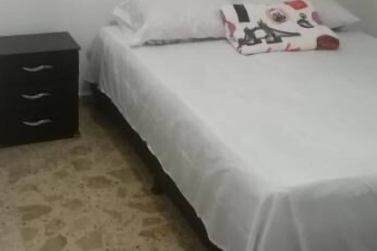 A small, sparsely furnished room with a single bed, a nightstand, and a wall-mounted shelf.