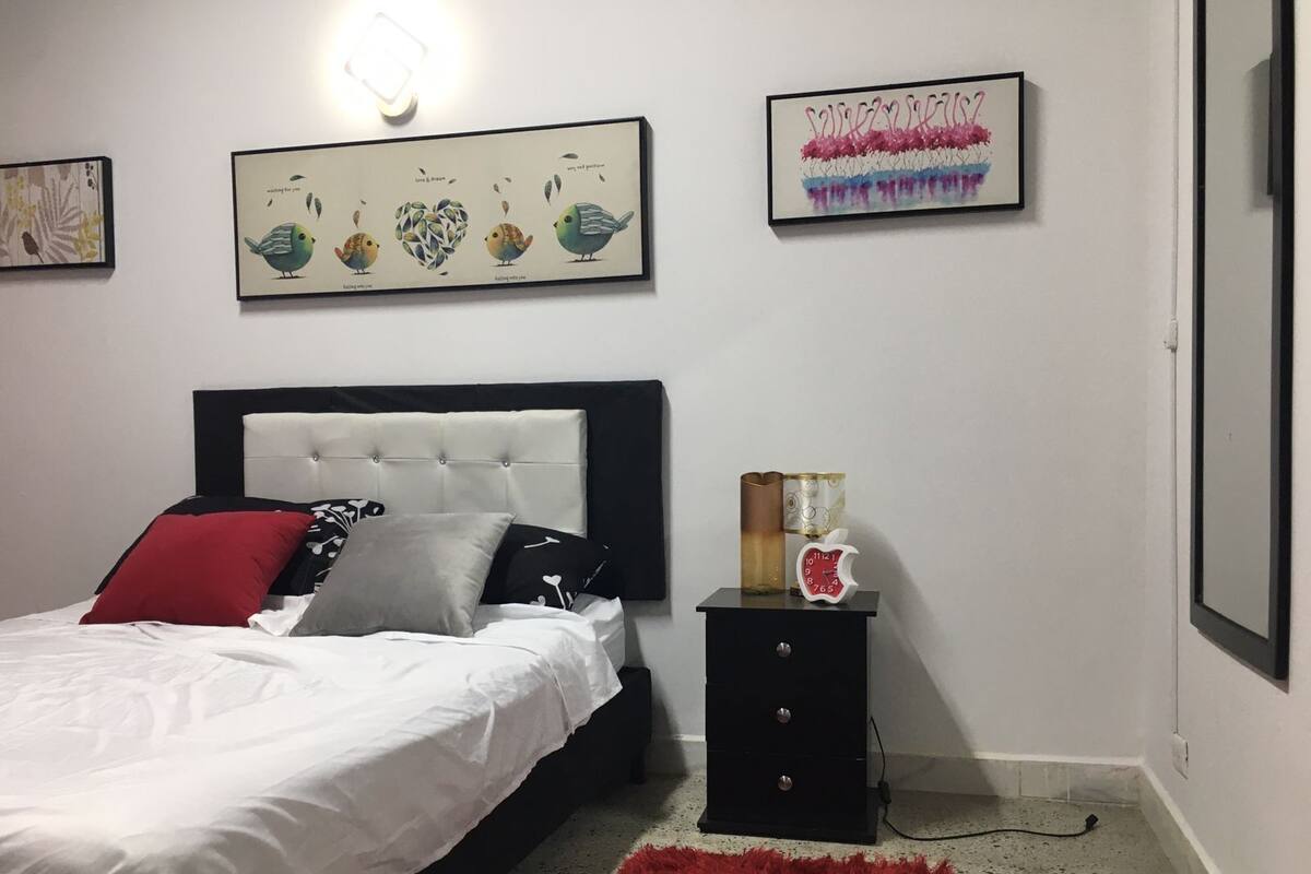 A bedroom with a bed, a nightstand, a red rug, and three framed artworks on the wall.