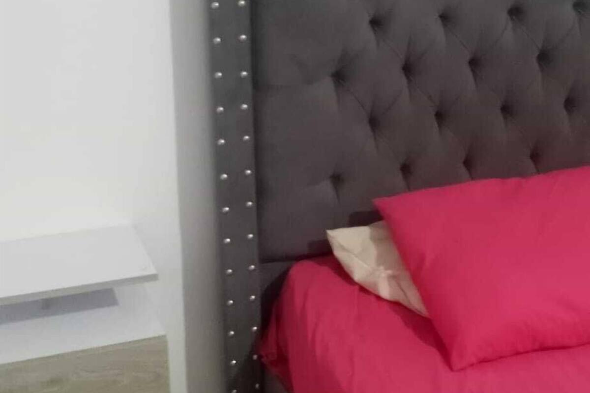 A bedroom with a tufted headboard, a red bedspread, a bedside table, and a drawer unit.