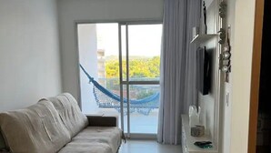 A modern hotel room with a sofa, a TV mounted on the wall, a small table, and a view of a blue sky and greenery outside. - Beautiful apartment in Bacutia (Guarapari)