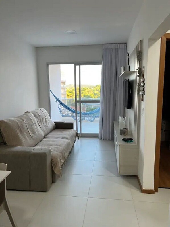 A modern hotel room with a sofa, a TV mounted on the wall, a small table, and a view of a blue sky and greenery outside.
