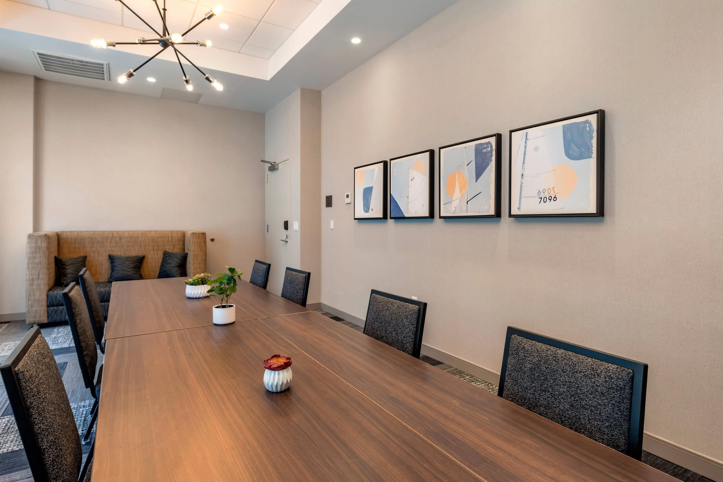 A conference room with a long wooden table, chairs, a plant, and framed artwork on the wall.