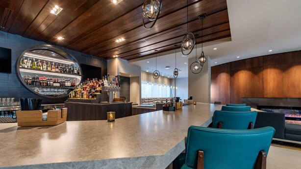 Un área de bar moderna con una barra curva, asientos con sillas color turquesa y una variedad de botellas y vasos en estantes.
