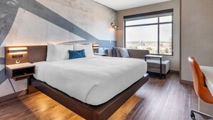 Habitación de hotel moderna con una cama grande, mesitas de noche con lámparas, un escritorio y vistas a la ciudad a través de un amplio ventanal.