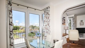 A dining area with a glass table and white chairs, a TV, and a view of a river and houses. - Harbour View (Pwllheli)
