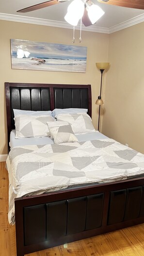 A bedroom with a wooden bed, a ceiling fan, a painting of a beach scene, and a floor lamp. - Travel light long-term (safe & relaxing )special discount for travel nurse (Veazie)