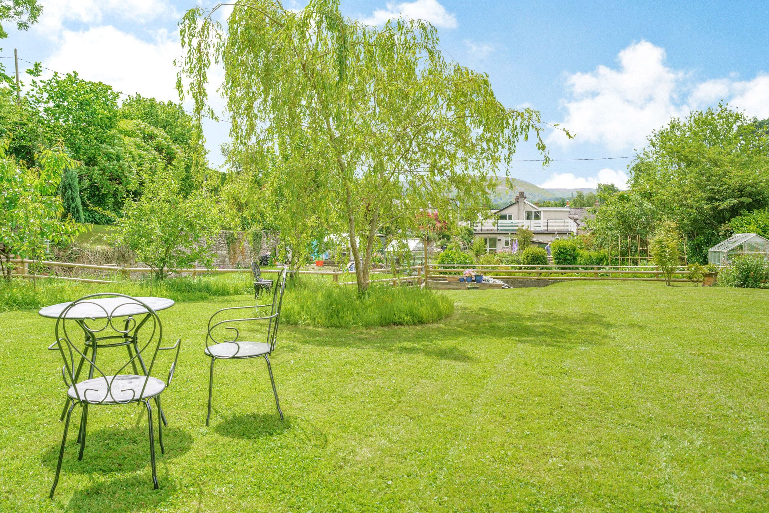Un espace extérieur verdoyant, avec des chaises et une table en métal, entouré d’arbres et avec une maison en arrière-plan.