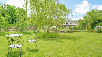 Un espace extérieur verdoyant, avec des chaises et une table en métal, entouré d’arbres et avec une maison en arrière-plan.