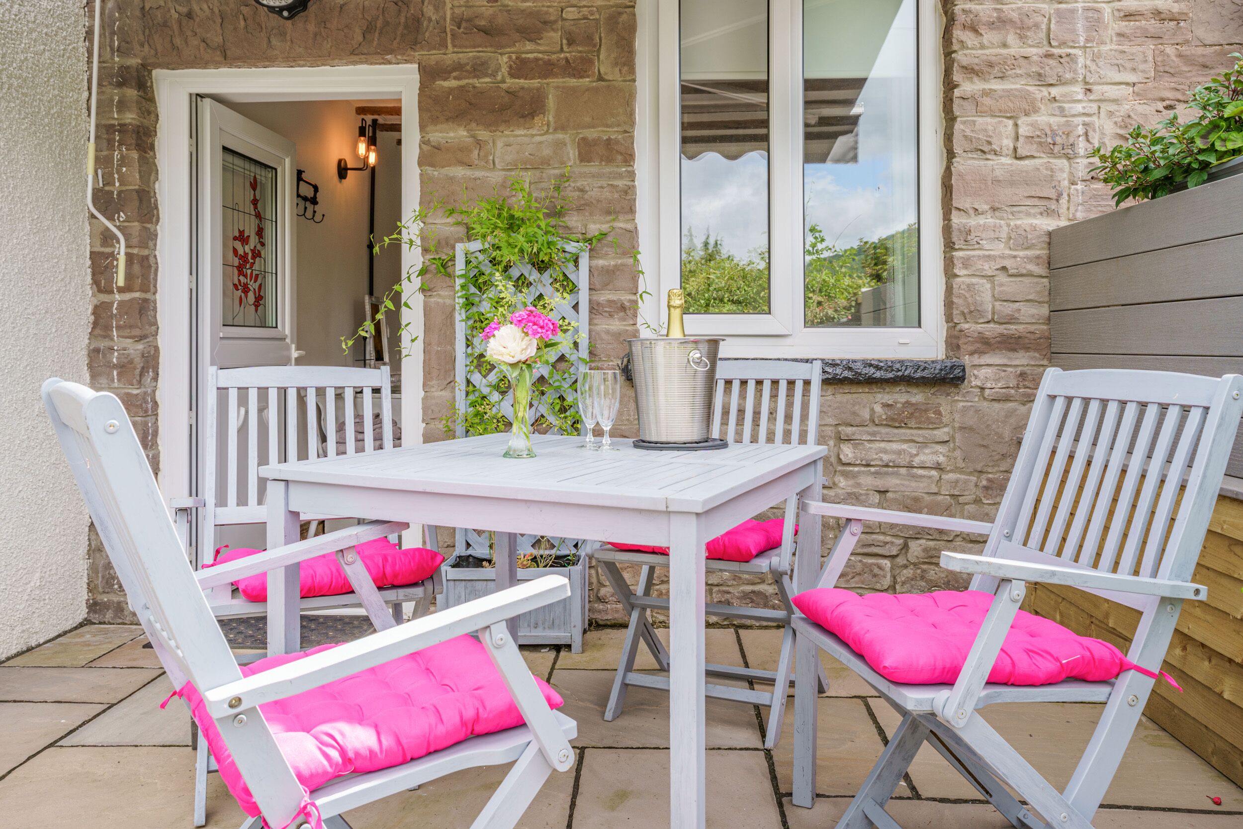 A patio with a white table and chairs, a vase with flowers, and a champagne bucket.