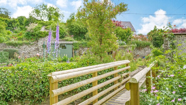 Un puente de madera en un jardín exuberante, con muros de piedra y diversas plantas y flores.