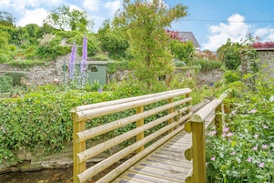 Un puente de madera en un jardín exuberante, con muros de piedra y diversas plantas y flores.