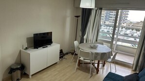 Un soggiorno moderno con una TV a schermo piatto su un mobile bianco, un tavolo da pranzo con sedie, un piccolo cestino e un'ampia finestra con vista su un porto turistico e degli edifici.