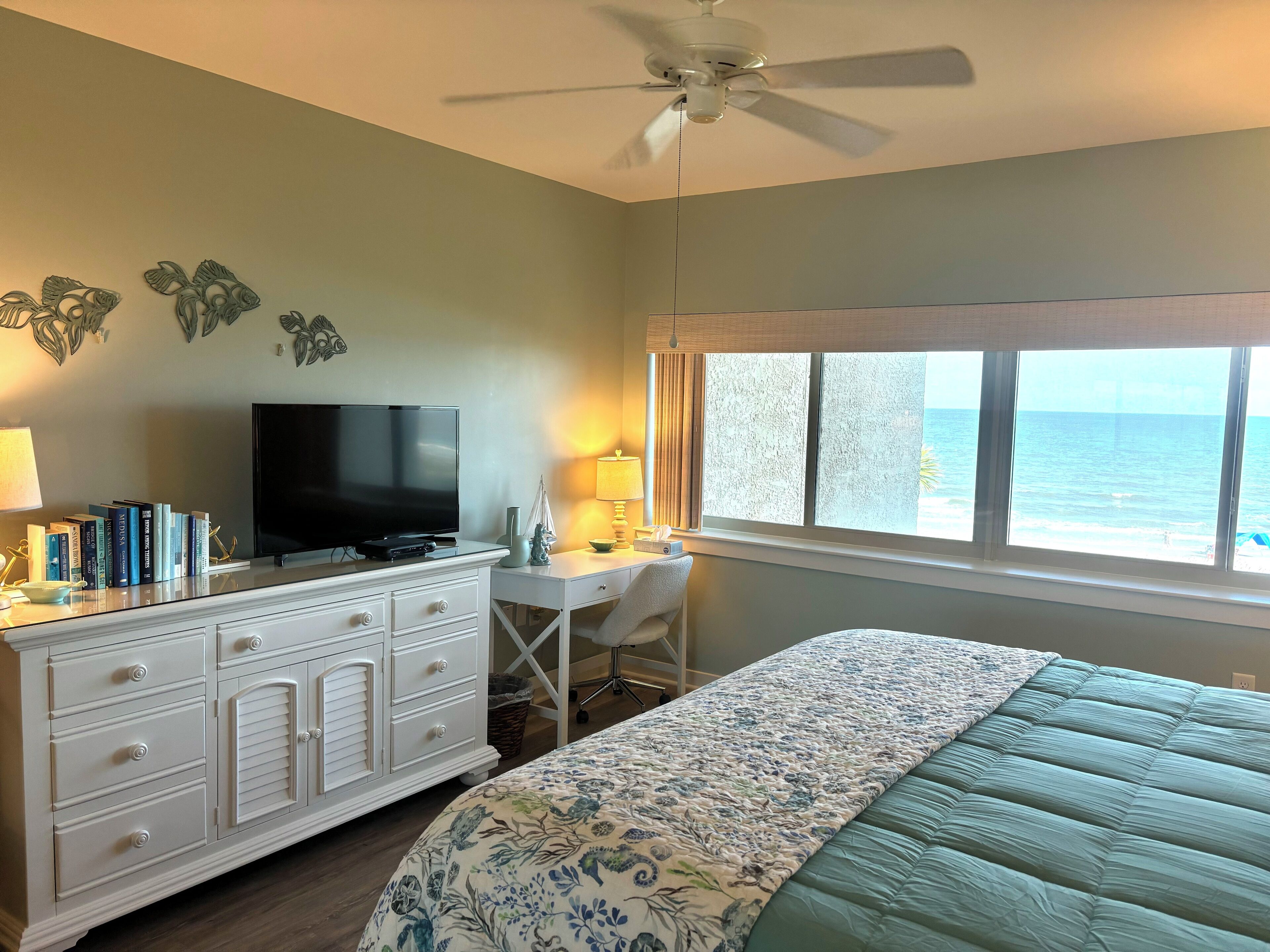 A bedroom with a bed, a TV on a dresser, a desk with a chair, and a window with a view of the ocean.